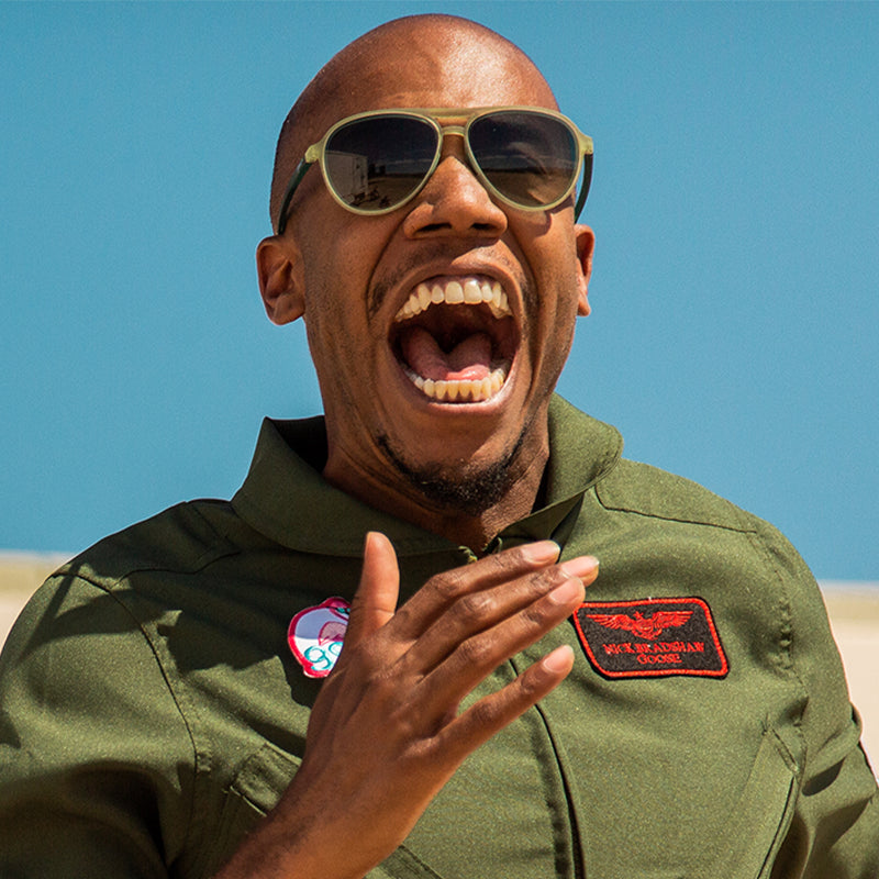 Ein Mann in einem grünen Militäroverall und einer kadettgrünen Pilotensonnenbrille mit grünen Verlaufsgläsern ruft fröhlich.