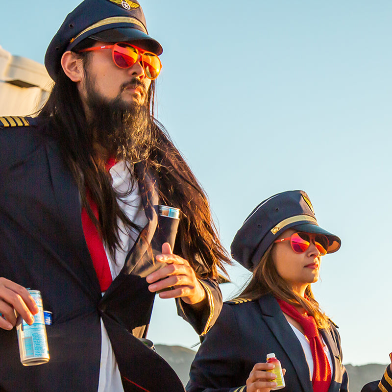 Zwei zerzauste Piloten mit roten Fliegersonnenbrillen und roten Gläsern steigen aus einem Flugzeug, Energydrinks in den Taschen.