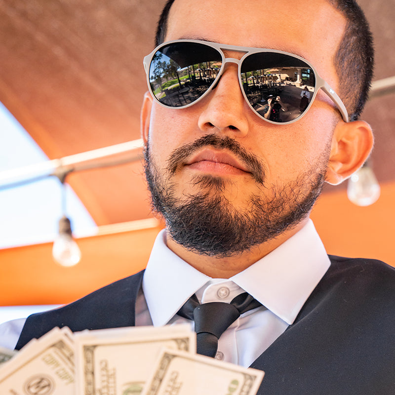 Ein Mann mit Krawatte und Weste in einem Golfclubhaus trägt eine graue Pilotensonnenbrille mit schwarzen Gläsern und fächelt sich mit Bargeld zu.