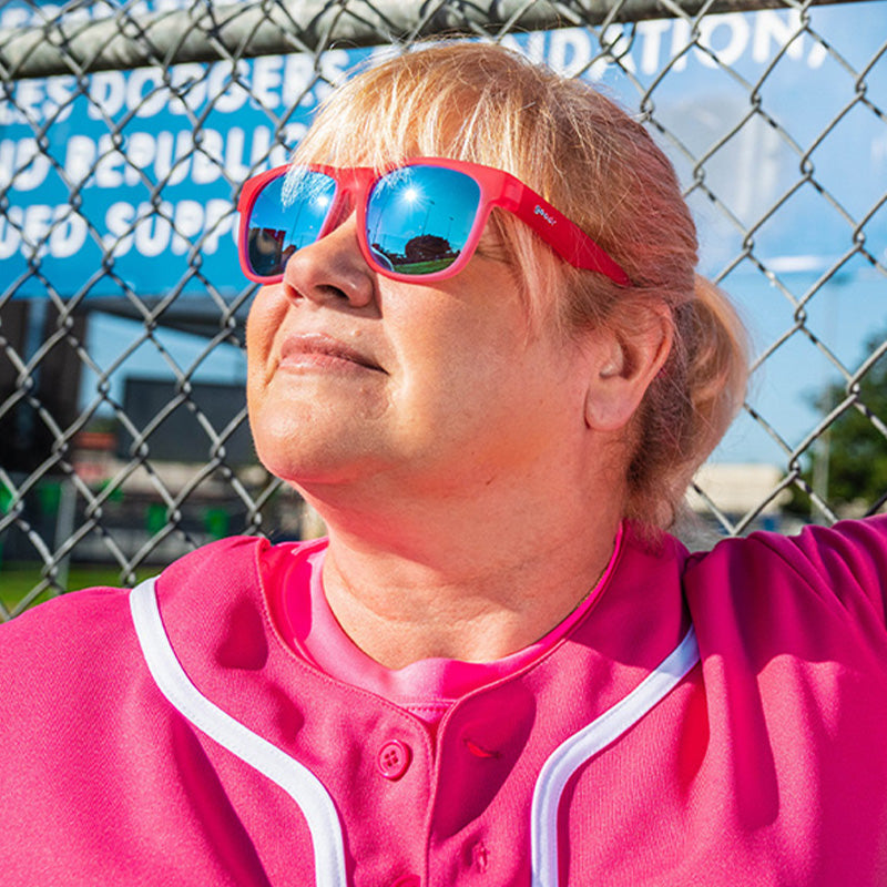 Eine Frau in einem pinkfarbenen Baseballtrikot und einer pinkfarbenen Sonnenbrille mit blauen Gläsern lehnt in einem Sonnenstrahl an einem Zaun.