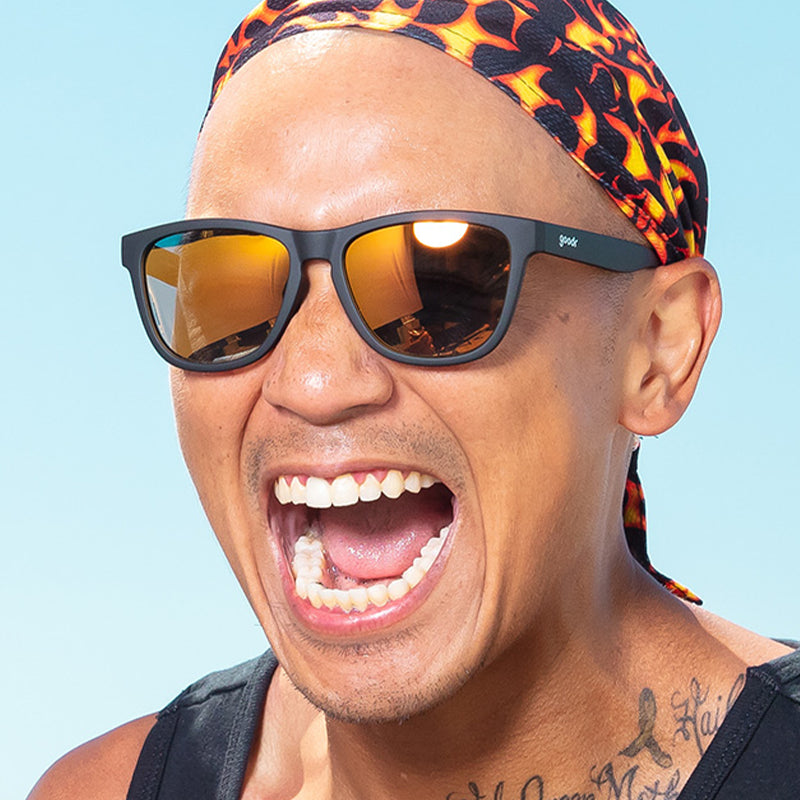 Ein Mann, der eine mit Flammen gemusterte Kutte und eine schwarze Sonnenbrille mit bernsteinfarbenen, reflektierenden Gläsern trägt, schreit fröhlich.