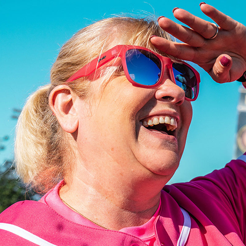 Dreiviertelansicht einer Dame in einem rosa Baseballdress und einer rosa Sonnenbrille mit blauen Gläsern, die auf einem Baseballfeld lächelt.