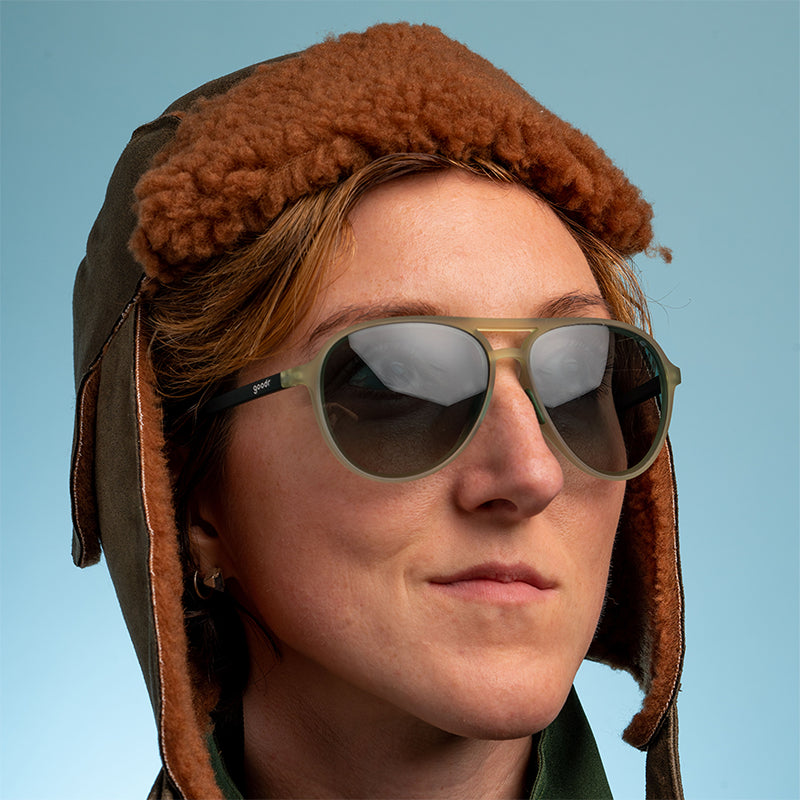 Eine Frau, die eine braune, wattierte Fliegerkappe trägt, schaut selbstbewusst aus dem Fenster und trägt eine kadettgrüne Fliegerbrille mit grünen Verlaufsgläsern.