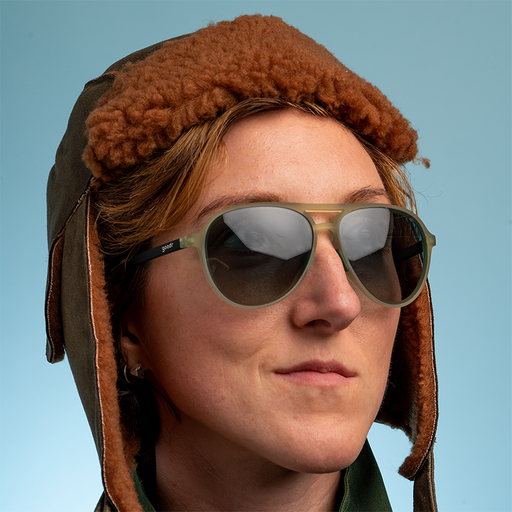 Eine Frau, die eine braune, wattierte Fliegerkappe trägt, schaut selbstbewusst aus dem Fenster und trägt eine kadettgrüne Fliegerbrille mit grünen Verlaufsgläsern.