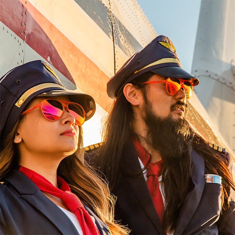 Zwei zerzauste Piloten mit roten Fliegersonnenbrillen mit roten Gläsern lehnen an einem Flugzeug und schauen in den Sonnenuntergang.