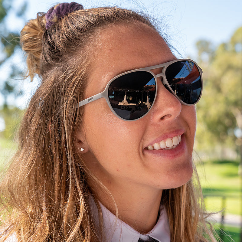 Eine lächelnde Frau mit einem Hemd mit Kragen in einem Golfclubhaus trägt eine graue Pilotensonnenbrille mit nicht reflektierenden Gläsern.