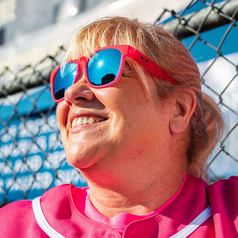 Eine Frau in einem rosafarbenen Baseballtrikot und einer rosafarbenen Sonnenbrille mit blau reflektierenden Gläsern lehnt lächelnd an einem Zaun.