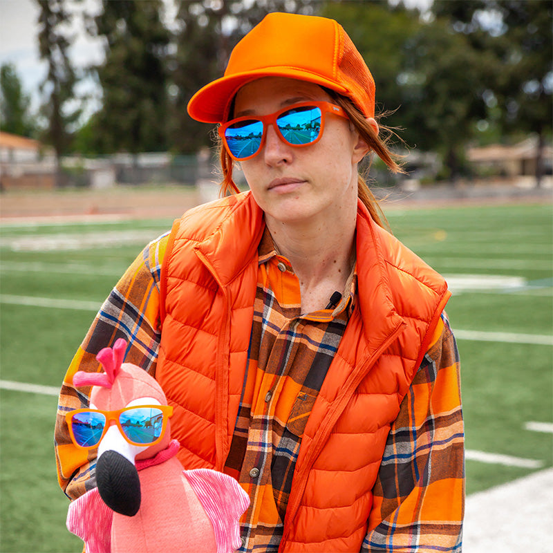 Eine Frau in einem leuchtend orangefarbenen Outfit hält einen ausgestopften rosa Flamingo, beide tragen eine orangefarbene Sonnenbrille mit blauen Gläsern.