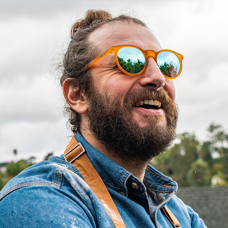 Ein bärtiger Mann mit Dutt lächelt, trägt eine runde orangefarbene Sonnenbrille mit hellblauen Gläsern und eine mit Mehl bedeckte Schürze.