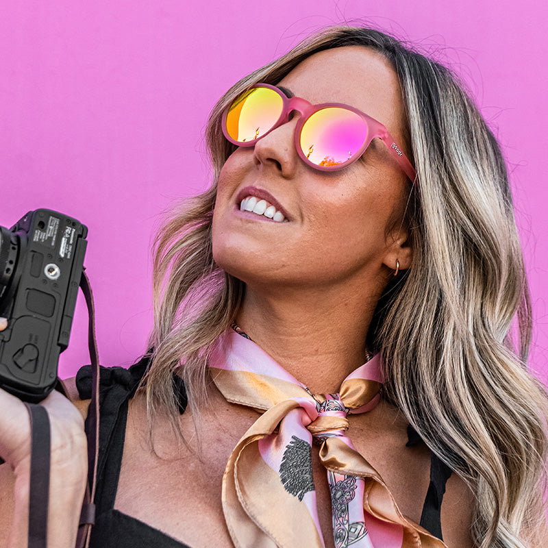 Eine elegante Frau mit einer runden rosa Sonnenbrille und einer Kamera in der Hand blickt zur Seite, hinter ihr eine pinkfarbene Wand.