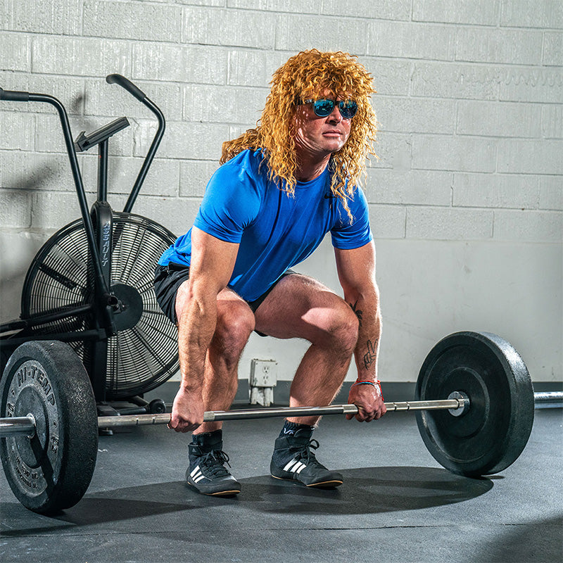 Ein Mann mit einer großen lockigen braunen Vokuhila-Perücke und einer schwarzen Sonnenbrille mit blauen reflektierenden Gläsern hebt eine schwere Hantel in einem Fitnessstudio.