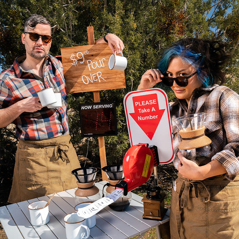 Zwei Hipster-Baristas mit runden braunen Sonnenbrillen und braunen Gläsern posieren an einem handwerklich gefertigten Pour-over-Kaffeestand.