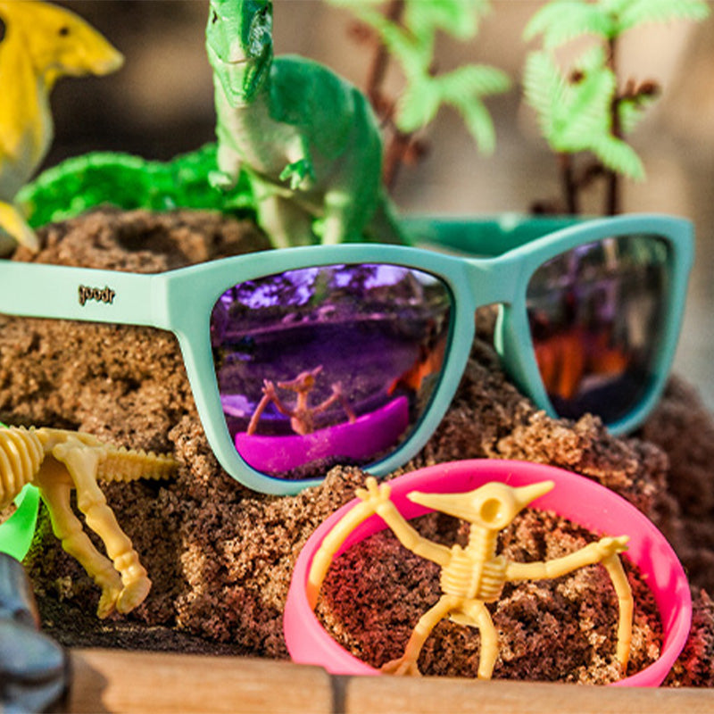 Dreiviertelansicht einer babyblauen Sonnenbrille mit lila reflektierenden Gläsern, die auf einem Erdhügel neben Dinosaurierspielzeug sitzt.