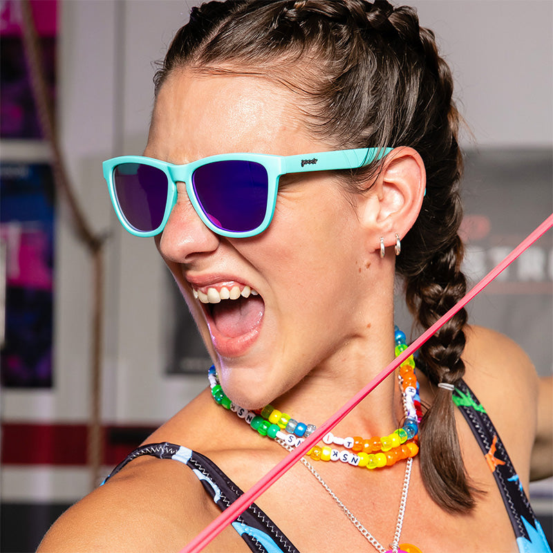 Eine Frau mit regenbogenfarbenen Plastikketten und einer babyblauen Sonnenbrille mit violetten, reflektierenden Gläsern grunzt in einem Fitnessstudio heftig.