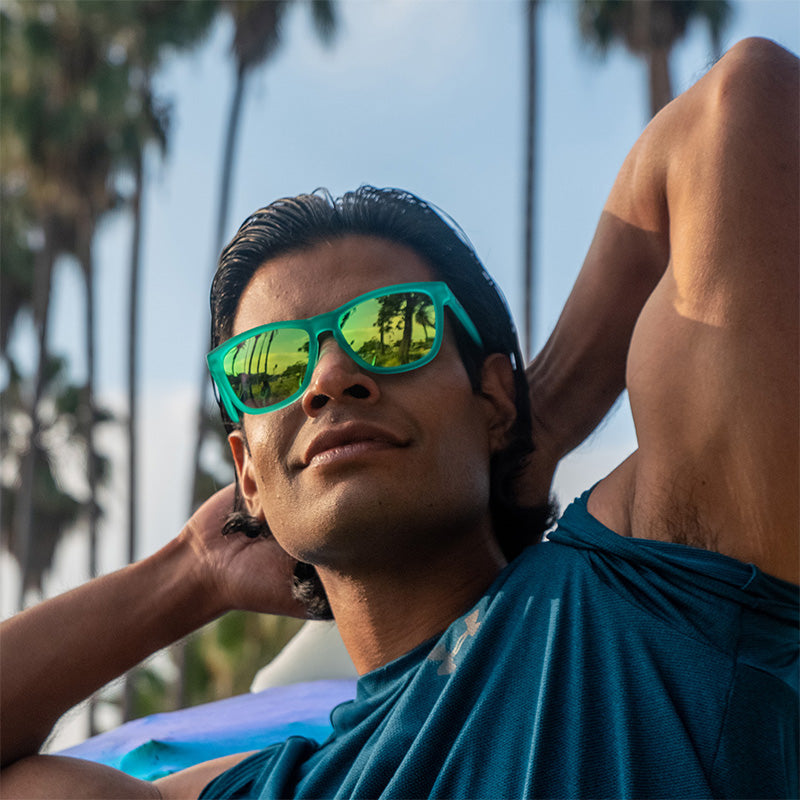 Ein grinsender, muskulöser Mann trägt eine blaugrüne Sonnenbrille mit blaugrünen, verspiegelten Gläsern und lässt seinen Bizeps spielen, während in der Ferne Palmen zu sehen sind.