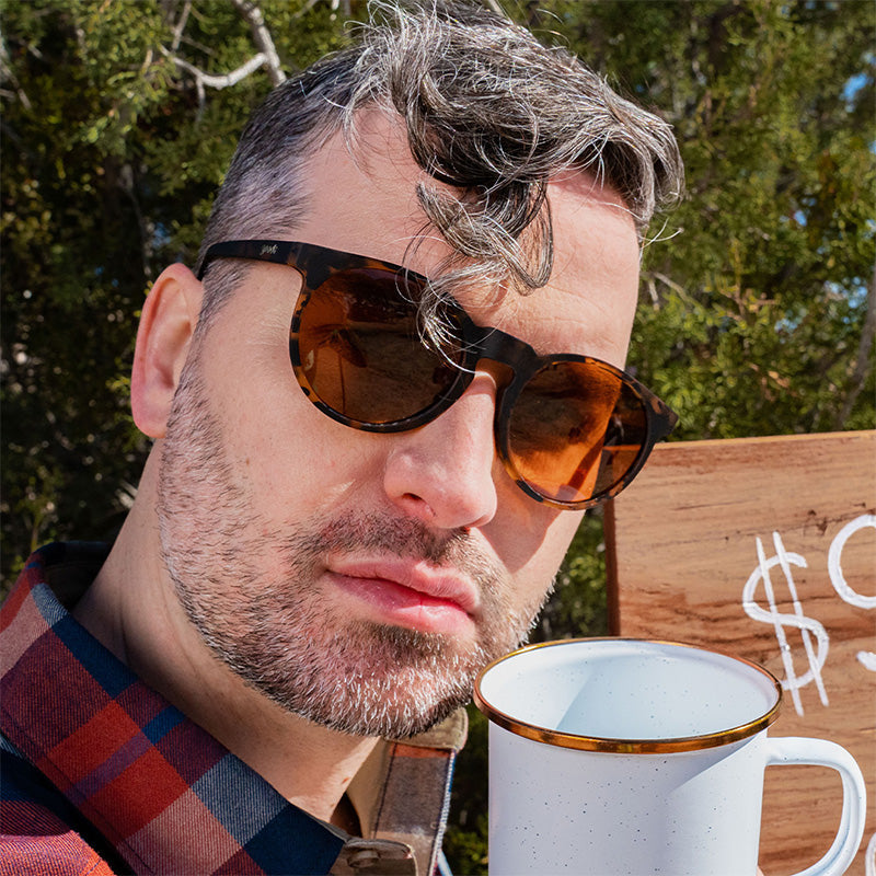 Ein Hipster-Barista blickt urteilend vor sich hin und trägt eine braune Schildpatt-Sonnenbrille mit braunen, nicht verspiegelten Gläsern.