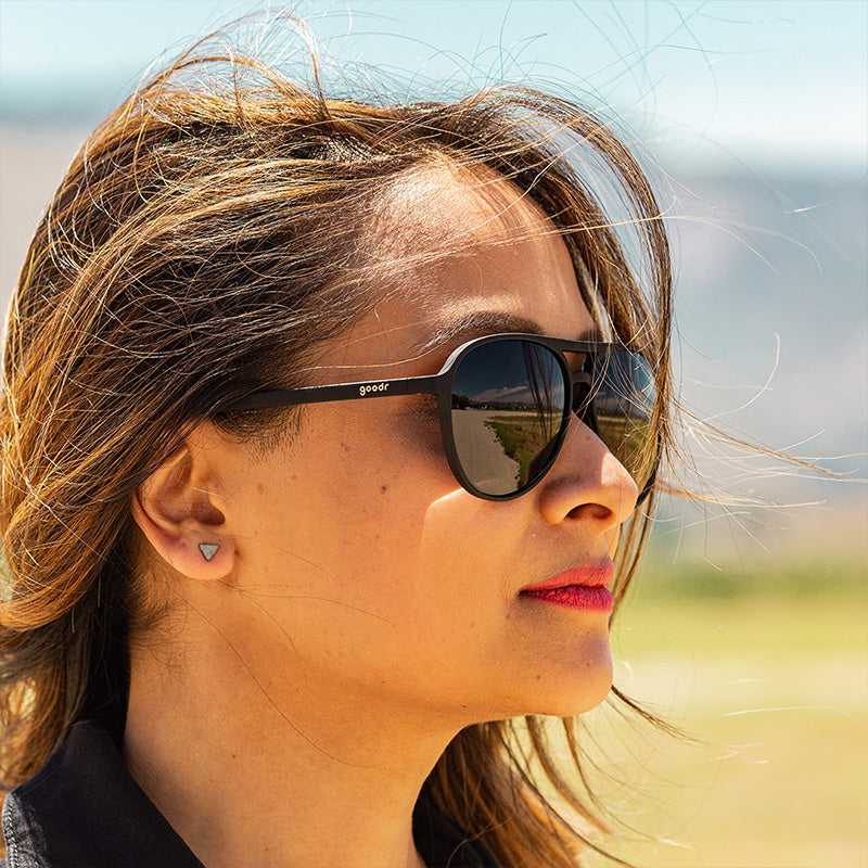 Eine Frau in einem schwarzen Overall und einer schwarzen Piloten-Sonnenbrille mit schwarzen, nicht verspiegelten Gläsern blickt grimmig nach vorne.