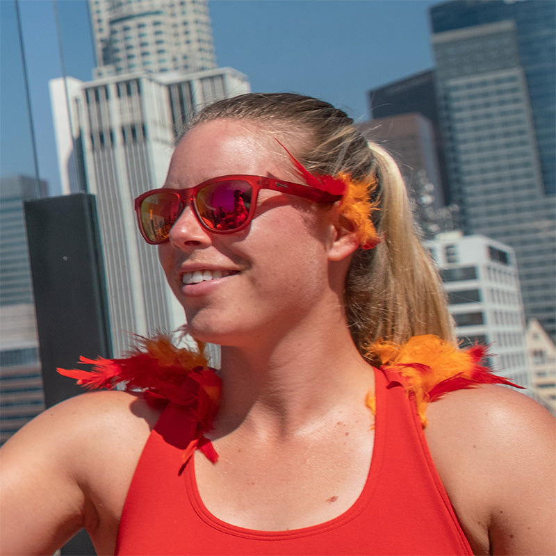 Eine Frau trägt eine rote Sonnenbrille mit roten Gläsern mit roten und orangefarbenen Federakzenten, im Hintergrund die Skyline einer Stadt.