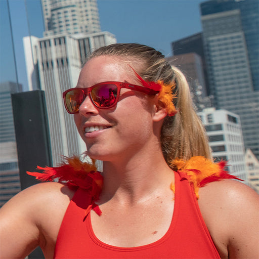 Eine Frau trägt eine rote Sonnenbrille mit roten Gläsern mit roten und orangefarbenen Federakzenten, im Hintergrund die Skyline einer Stadt.